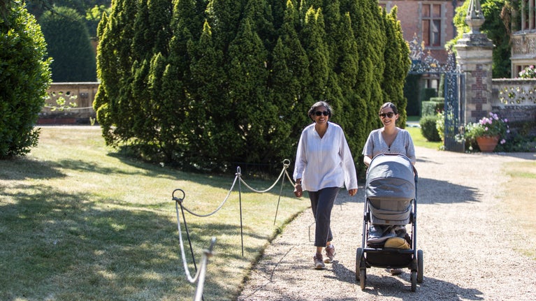 A multi generational family taking a leisurely stroll along a shady path in the gardens on a bright, hot sunny summer's day. A mature daughter is talking to her mother (grandmother) who is pushing a pram.
